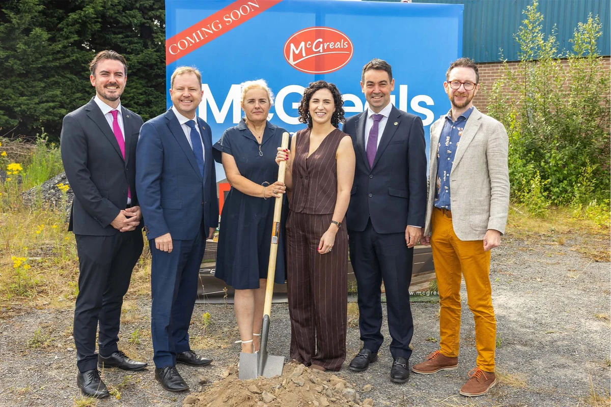 A group gathered with a ceremonial spade in front of a McGreals coming-soon sign in Jigginstown